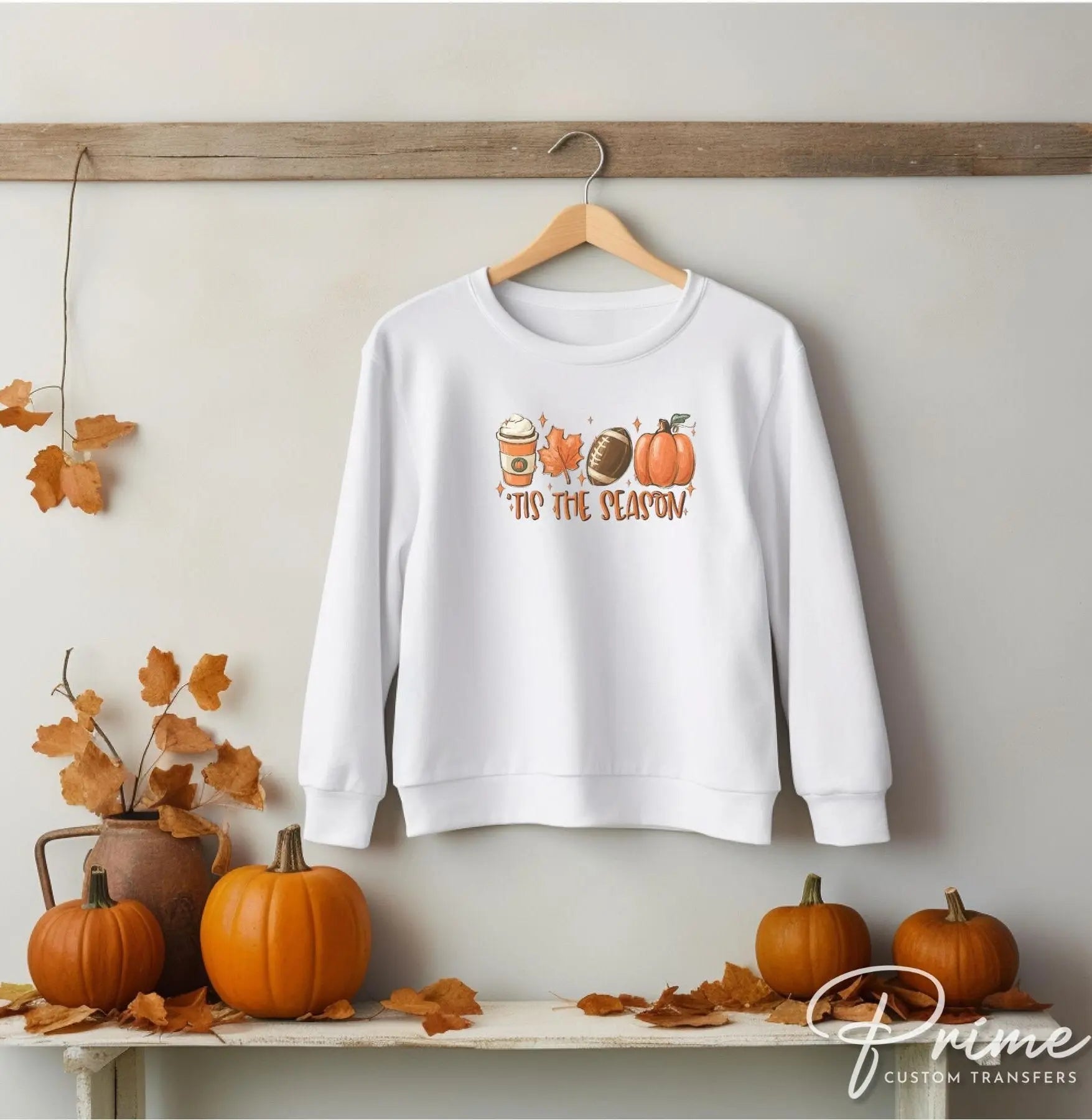 A picture of a white sweatshirt with cute pictures of coffee, leaves, footballs and pumpkins and the words Tis The Season. DTF transfers, DIY , Make your own shirt
